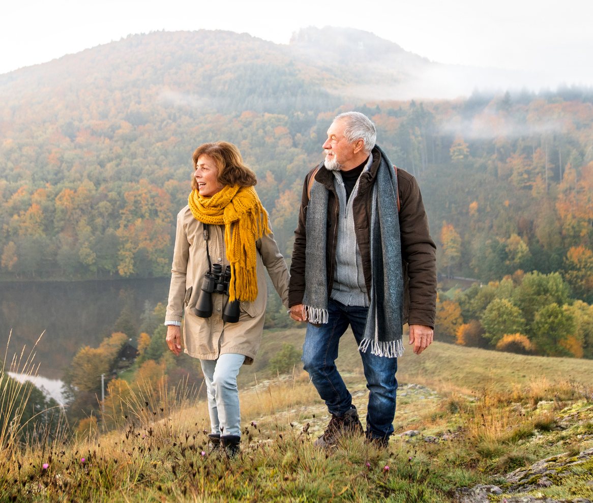A retired couple walking hand in hand on a hillside above autumn woods